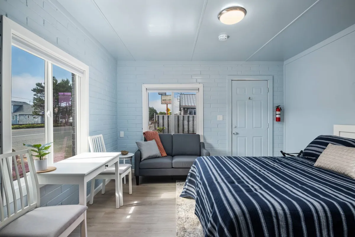 A cozy interior featuring a bed with striped bedding, a cushioned sofa, a small dining table with chairs, and a window providing an outdoor view. The space is well-lit and thoughtfully arranged for both relaxation and socializing, highlighting the recent upgrades.