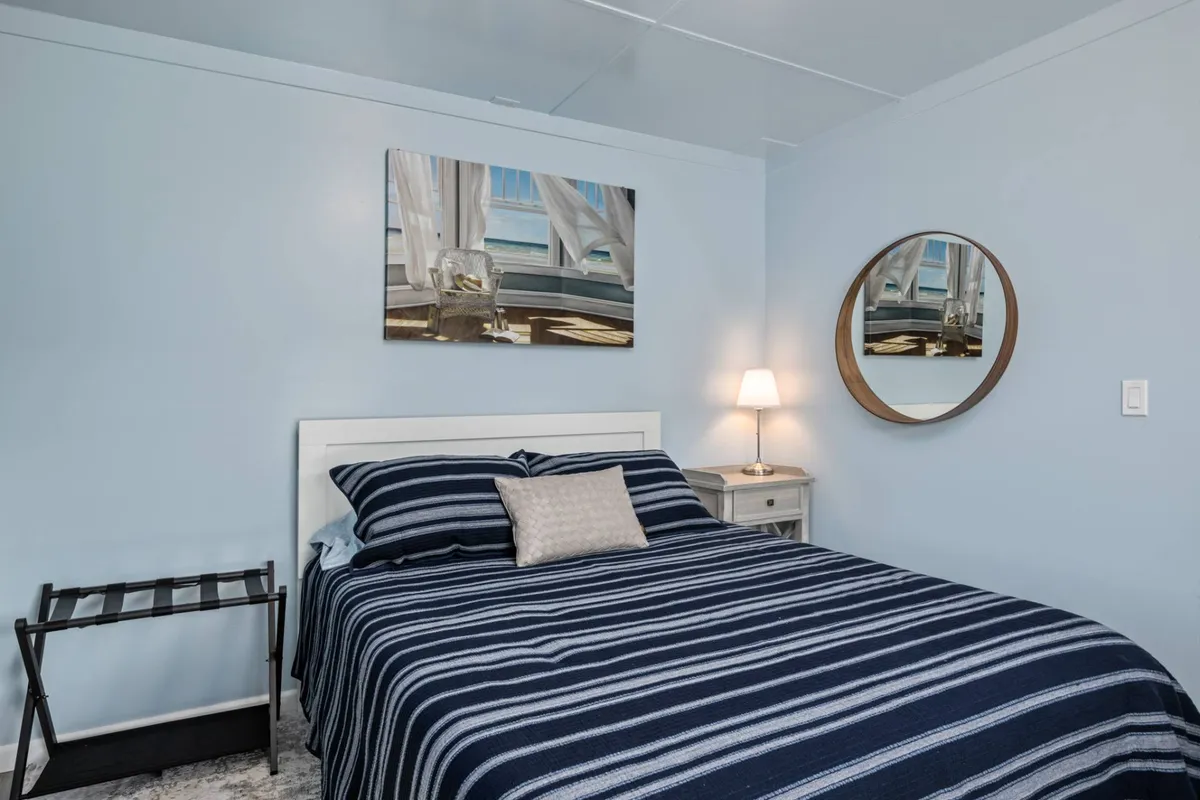 A tranquil bedroom scene with striped bedding, a round mirror, framed artwork, and a nightstand with a lamp. Soft, natural light enters the room, highlighting the fresh, recently renovated feel and comfortable, serene vibe.