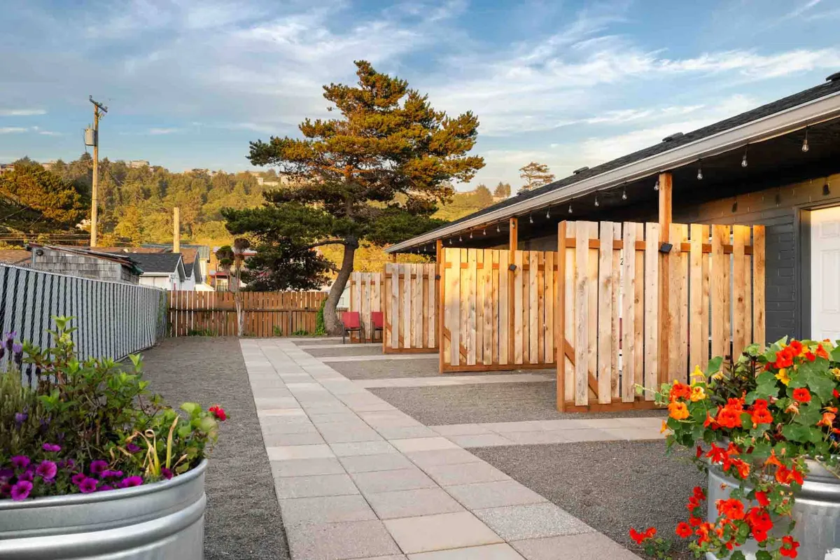A welcoming garden walkway bordered by wooden fencing and flower pots, with people walking and relaxing in the sunset light. The area exudes warmth and is an ideal setting for both human and pet relaxation after a day of adventure.