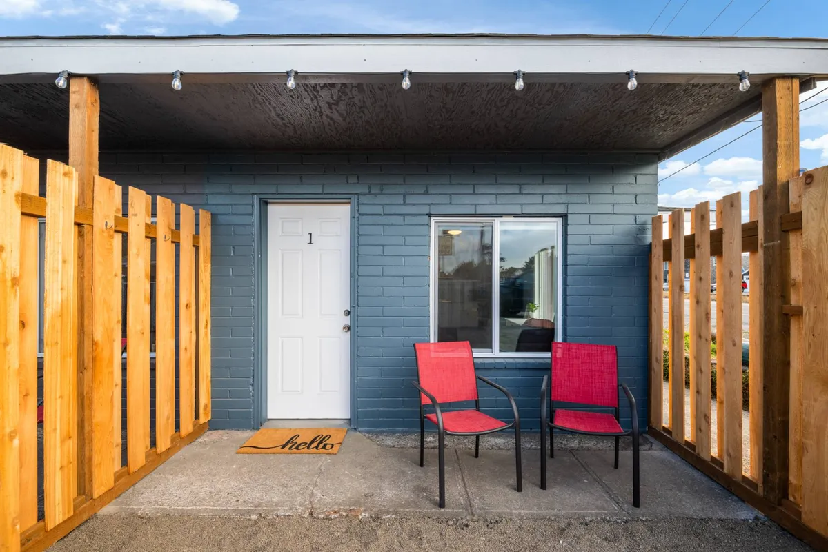 This image shows a charming cottage entrance with a front door, a welcome mat, and a semi-private outdoor seating area bordered by a fence. The arrangement highlights the cozy, personal patio space that is just steps from the living area, ideal for unwinding or enjoying a quiet morning outside.