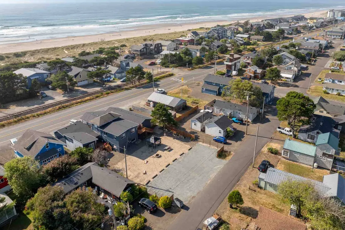 A wide exterior view encompassing the sandy beach, a nearby road, houses, and trees. The scene features people strolling along the beach, with the ocean and sunset in the background, evoking the relaxed, social atmosphere of a coastal community.