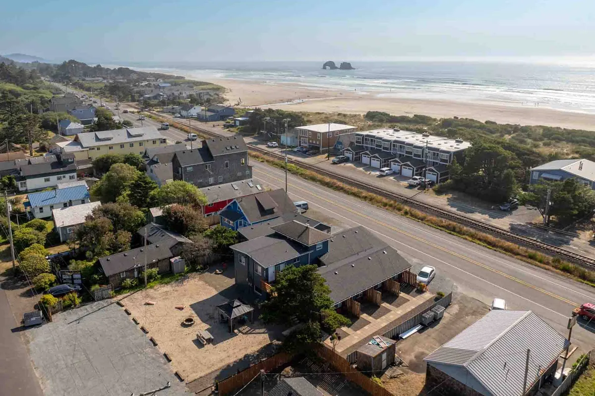 A panoramic exterior shot of the classic Rockaway Beach cottages lined up near the beach, with the ocean and dramatic rock formations in the distance. The scene bustles with activity—people walking, preparing for beach outings, or heading out to explore, creating a vibrant, lively mood synonymous with beach vacations.