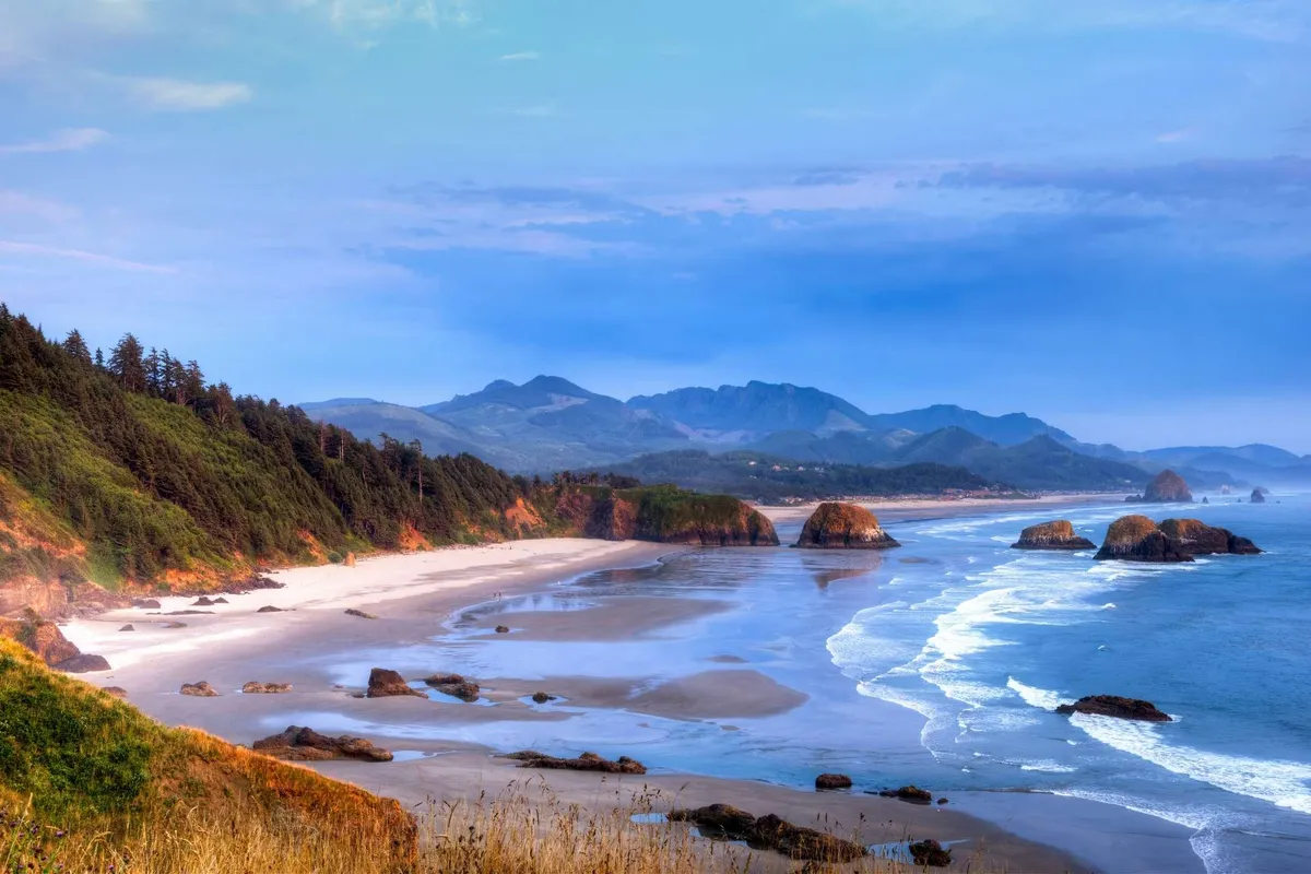 The scene presents a breathtaking coastal landscape with rocky formations, a sandy beach, rolling ocean waves, and distant mountains under a tranquil sky. The image evokes the natural beauty and adventure awaiting just minutes from the property.