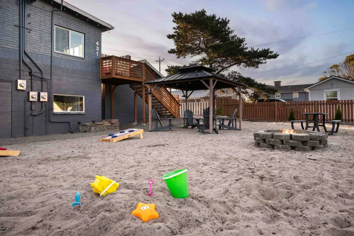 This exterior view centers on a cheerful gazebo area with seating, a sandbox for children, a fire pit, and stairs leading up to additional spaces. The gazebo is the focal point and is clearly set up for community gatherings, group meals, or relaxed socializing.